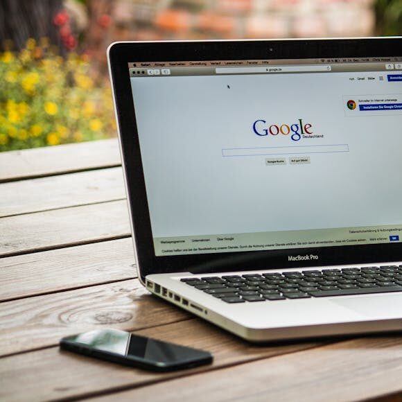 A MacBook Pro displaying Google Search on a wooden table outdoors, next to a smartphone.